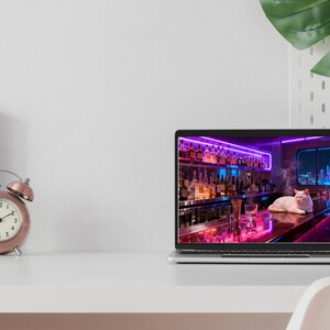 May include: A rose gold alarm clock with a white face sits next to a laptop displaying a neon-lit bar scene with a white cat. The laptop is open on a white desk. A green plant is in the background.