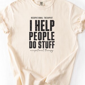 May include: A cream-coloured short-sleeve t-shirt featuring black text that reads OCCUPATIONAL THERAPIST, I HELP PEOPLE DO STUFF, and occupational therapy in a script font. The shirt has a crew neckline and a Comfort Colors brand label.