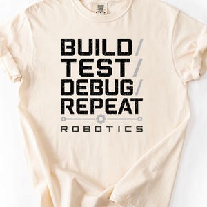 Op de afbeelding: Een crèmekleurig t-shirt met korte mouwen en zwarte, verweerde tekst die BUILD / TEST / DEBUG / REPEAT leest, met een grijs tandwielpictogram en het woord ROBOTICS eronder, gecentreerd op de borst van het kledingstuk.