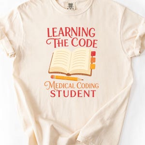 May include: A cream-colored t-shirt featuring a graphic of an open book with three colorful tabs, a yellow pencil, and the red and orange text "LEARNING THE CODE" and "MEDICAL CODING STUDENT" printed in a distressed, vintage-style font on the front.