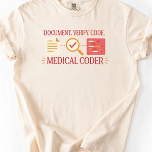 Puede incluir: Una camiseta de manga corta color crema con un gráfico que incluye el texto "DOCUMENT. VERIFY. CODE." y "MEDICAL CODER.". El diseño presenta un icono de documento, una lupa con una marca de verificación y un bloque de código rojo.