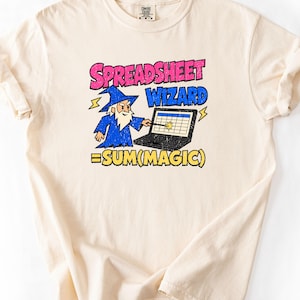 May include: A cream-colored t-shirt featuring a graphic of a wizard in a blue robe pointing a wand at a laptop screen displaying a spreadsheet. The text reads SPREADSHEET WIZARD and =SUM(MAGIC) in pink, blue, and yellow with lightning bolt accents.