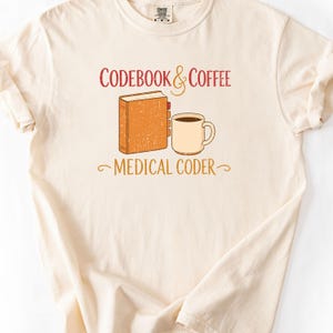 Op de afbeelding: Een crèmekleurig t-shirt met korte mouwen en een afbeelding van een bruin boek en een beige koffiemok. De tekst CODEBOOK & COFFEE staat boven de afbeelding en MEDICAL CODER eronder in een verweerd rood en goudkleurig lettertype.