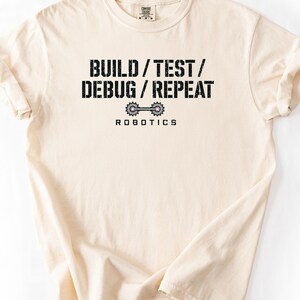 May include: A cream-colored short-sleeve t-shirt featuring black distressed text that reads BUILD / TEST / DEBUG / REPEAT. Below the text is a graphic of two connected grey gears with pink centers, followed by the word ROBOTICS in black capital letters.