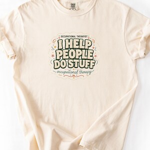 May include: A cream-coloured short-sleeve t-shirt featuring a central graphic with the text "OCCUPATIONAL THERAPIST I HELP PEOPLE DO STUFF occupational therapy" in dark green and beige, surrounded by small red and blue stars and decorative lines.