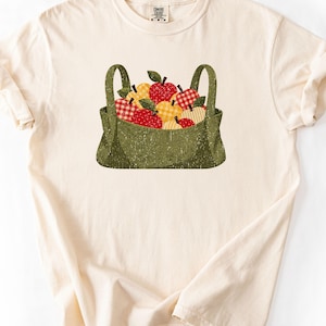 Könnte beinhalten: Ein cremefarbenes Kurzarm-T-Shirt mit einer zentralen Grafik einer abgenutzten olivgrünen Tragetasche, die mit verschiedenen Äpfeln gefüllt ist. Die Äpfel zeigen rote und gelbe Muster, darunter Vichy-Karos, Punkte und einfarbige Designs.