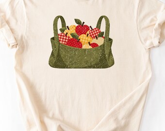 Puede incluir: Una camiseta de manga corta color crema con un gráfico central de una bolsa de tela verde oliva desgastada llena de manzanas. Las manzanas muestran patrones rojos y amarillos, incluyendo cuadros vichy, lunares y colores sólidos con tallos marrones.