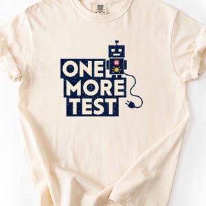 Może przedstawiać: Kremowy t-shirt z krótkim rękawem z ciemnoniebieską grafiką i napisem ONE MORE TEST. Wzór przedstawia małego robota z czerwono-żółtym okrągłym detalem na klatce piersiowej oraz czarny przewód zasilający zakończony wtyczką.