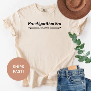 May include: Cream-colored t-shirt with the text "Pre-Algorithm Era" and "Est. 2016" printed in black. The shirt is paired with blue jeans and a brown hat. A round sticker with the text "SHIPS FAST!" is also visible.