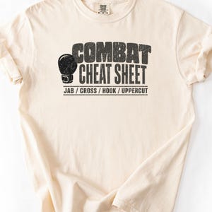 May include: A cream-colored short-sleeve t-shirt featuring a black graphic print. The design includes a boxing glove icon, the text "COMBAT CHEAT SHEET" in a distressed font, and the words "JAB / CROSS / HOOK / UPPERCUT" underlined below.