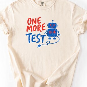 Può includere: Una t-shirt a maniche corte color crema con una stampa grafica che presenta il testo rosso "ONE MORE" sopra il testo blu "TEST". Accanto al testo c'è l'illustrazione di un robot blu con occhi, bocca e un ingranaggio di colore rosso sul petto.