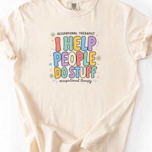 May include: A cream-coloured short-sleeve t-shirt featuring a graphic print with the text "OCCUPATIONAL THERAPIST I HELP PEOPLE DO STUFF occupational therapy" in colourful, distressed bubble letters surrounded by small flower and star icons.
