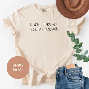 May include: Cream-colored t-shirt with the phrase "I WON'T TAKE NO FOR AN ANSWER" in black lettering. The shirt is paired with blue jeans and a brown hat. The shirt is a casual wear item.