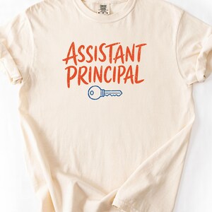 May include: A cream-colored short-sleeve t-shirt featuring the text "ASSISTANT PRINCIPAL" printed in a distressed orange font. Below the text is a simple blue graphic of a key. The shirt has a crew neckline and a standard fit.
