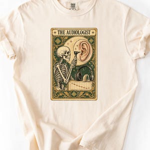 Puede incluir: Una camiseta color crema con un gráfico rectangular de borde verde y dorado. El diseño muestra un esqueleto usando un otoscopio en un oído humano, junto a auriculares, un audífono y un gráfico de líneas descendente. El texto dice THE AUDIOLOGIST.
