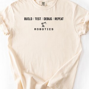 Peut inclure: Un t-shirt à manches courtes de couleur crème avec un texte noir indiquant BUILD / TEST / DEBUG / REPEAT, un petit graphique d'un bras robotique au centre et le mot ROBOTICS imprimé en lettres capitales noires sous le graphique.