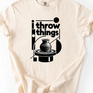 May include: A cream-colored short-sleeve t-shirt featuring a black graphic print. The design includes the text "i throw things" above an illustration of a pottery vase on a spinning wheel, surrounded by geometric shapes and lines.