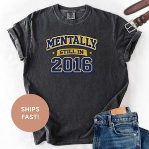 Puede incluir: Camiseta gris oscuro con el texto "MENTALLY STILL IN 2016" en azul y amarillo. La camiseta tiene un aspecto vintage. Un cinturón marrón y unos vaqueros azules son visibles en el fondo.