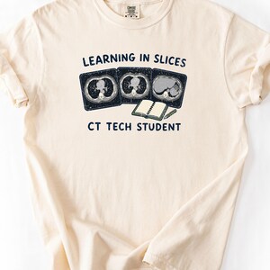 May include: A cream-colored short-sleeve t-shirt featuring a graphic with three CT scan images, an open notebook, and a pencil. The text reads "LEARNING IN SLICES" above the images and "CT TECH STUDENT" below, all printed in a dark navy blue font.