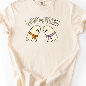 Puede incluir: Una camiseta de manga corta color crema con un gráfico de dos fantasmas frente a frente. Un fantasma lleva un cinturón naranja y el otro un cinturón morado. Sobre los fantasmas, el texto BOO-JITSU está impreso en una fuente oscura.