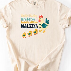 Może przedstawiać: Kremowy t-shirt z krótkim rękawem z grafiką przedstawiającą kurę i trzy pisklęta. Tekst brzmi Medical Coding Farm Edition Pecked by a Chicken W61.33XA. Wzór wykorzystuje żółte, turkusowe i czerwone kolory z efektem postarzenia materiału.