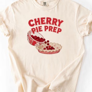 Könnte beinhalten: Ein cremefarbenes Kurzarm-T-Shirt mit einer Grafik, die eine Schüssel zeigt, aus der rote Kirschen in einen Tortenboden geschüttet werden. Der Text CHERRY PIE PREP ist darüber in einem roten Punktmuster gedruckt. Das Design nutzt rot-weiße Vichy-Muster.