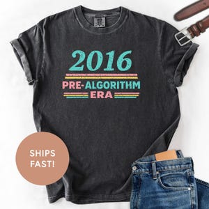 Puede incluir: Camiseta gris oscuro con el texto "2016 PRE-ALGORITHM ERA" en turquesa, rosa y amarillo. La camiseta tiene un aspecto vintage y se combina con vaqueros azules y un cinturón marrón.