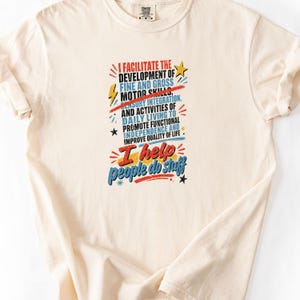 May include: A cream-coloured short-sleeve t-shirt featuring a graphic print with the text: I FACILITATE THE DEVELOPMENT OF FINE AND GROSS MOTOR SKILLS, SENSORY INTEGRATION, AND ACTIVITIES OF DAILY LIVING TO PROMOTE FUNCTIONAL INDEPENDENCE AND IMPROVE QUALITY OF LIFE. I help people do stuff. The design includes red, blue, and yellow accents with lightning bolts and stars.