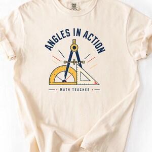 May include: A cream-colored short-sleeve t-shirt featuring a graphic with the text "ANGLES IN ACTION" and "MATH TEACHER". The design includes a dark blue drafting compass, a yellow protractor, and a yellow triangle ruler with red accents.