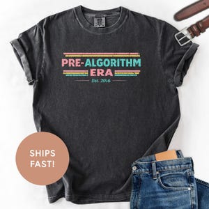 May include: Dark gray t-shirt with the text "PRE-ALGORITHM ERA" in pink, teal, and yellow stripes, with "Est. 2016" below. The shirt is paired with blue jeans and a brown belt.