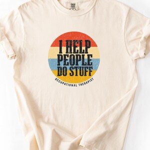 May include: A cream-coloured short-sleeve t-shirt featuring a circular graphic with horizontal stripes in red, cream, blue, and yellow. The text reads I HELP PEOPLE DO STUFF and OCCUPATIONAL THERAPIST in black, distressed, block-style lettering.
