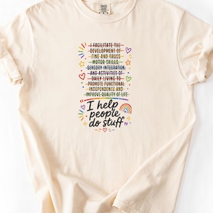 May include: A cream-coloured short-sleeve t-shirt featuring a graphic with rainbow-coloured lines crossing out text that reads: I FACILITATE THE DEVELOPMENT OF FINE AND GROSS MOTOR SKILLS, SENSORY INTEGRATION, AND ACTIVITIES OF DAILY LIVING TO PROMOTE FUNCTIONAL INDEPENDENCE AND IMPROVE QUALITY OF LIFE. Below this, the text reads: I help people do stuff, accompanied by small rainbow, star, and heart icons.