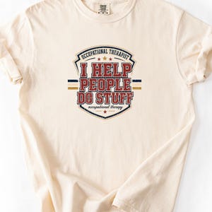 May include: A cream-coloured short-sleeve t-shirt featuring a distressed graphic design in red, blue, and gold. The text reads OCCUPATIONAL THERAPIST, I HELP PEOPLE DO STUFF, and occupational therapy, arranged within a shield-shaped emblem with stars.