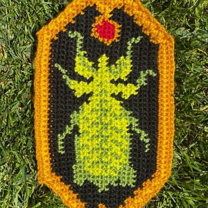 May include: A handmade crochet patch featuring a stylized insect design. The insect is primarily lime green, set against a black background, and framed by a gold border. A small red accent is at the top. The patch is placed on green grass.