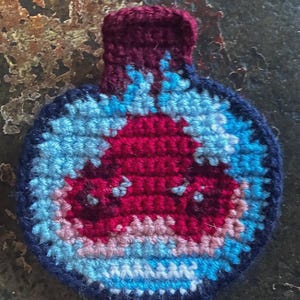 May include: Handmade crochet coaster in a bottle shape. The coaster features a dark blue border, light blue and pink accents, and a central red design. The top is a deep burgundy color.