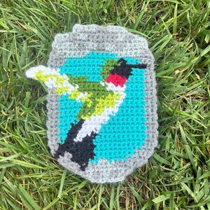 May include: A rectangular crochet patch featuring a central image of a hummingbird with green, white, and black feathers and a bright red throat. The bird is set against a vibrant turquoise background, all framed by a textured light gray border.