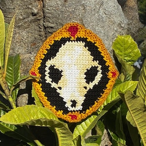 May include: A crocheted patch featuring a skull design. The patch has a black and white skull in the center, surrounded by a black border, a gold border, and red accents. The patch is displayed among green leaves and a tree trunk.