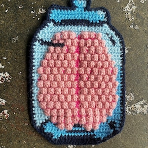 May include: A handmade crochet coaster featuring a pink brain inside a blue jar. The brain has a pink center line. The jar is outlined in dark blue and has a light blue and dark blue lid.