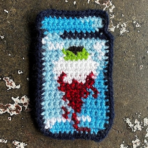 May include: A handmade crochet patch featuring a stylized eye design. The patch has a dark blue border, with a light blue and white background. The eye has a green iris, black pupil, and red veins.