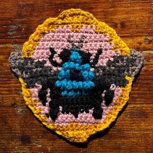 May include: A handmade crochet coaster with an insect design. The coaster has a yellow border, a pink and black body, and a blue head. It is placed on a wooden surface.