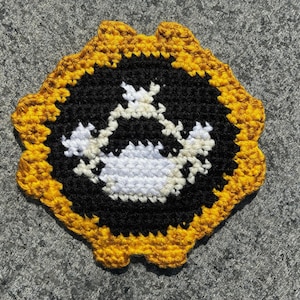 May include: A handmade crochet coaster featuring a black center with a white and cream-colored design, framed by a scalloped gold border. The coaster is round and appears to be made of yarn.