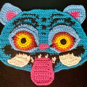 Könnte beinhalten: Eine handgefertigte Häkelmaske in Form eines Tigergesichts. Die Maske hat eine hellblaue Basis mit schwarzen, rosa, weißen, gelben und orangefarbenen Details. Der Tiger hat große, ausdrucksstarke Augen und eine rosa Zunge.