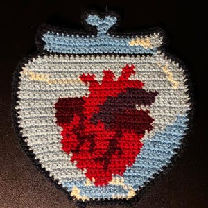 May include: A handmade crochet coaster featuring a red heart design inside a light blue jar shape. The jar has a black outline and a small heart detail at the top. The background is a neutral gray.