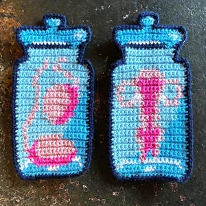 May include: Two handmade crochet jar-shaped coasters. Each coaster features a blue background with pink and white floral designs. The coasters have a dark blue border and a jar lid design at the top.