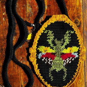 May include: A black crocheted crossbody bag with a yellow and black border. The bag features a green, red, yellow, and white insect design. The strap is black and the bag is on a wooden surface.