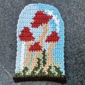 May include: A crocheted beanie featuring a mushroom design. The hat has a light blue background with red mushroom caps, tan stems, and green and brown foliage at the base. The brim is dark brown.