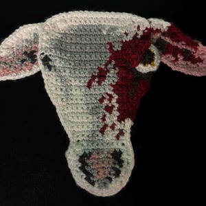 May include: Crocheted cow head in white, grey, and red. The cow head has pink and black details on the ears and muzzle. The red colour is splattered across the face. The cow head is on a black background.