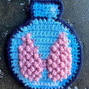May include: A handmade crochet patch in the shape of a blue bottle with a pink, textured design inside. The patch has a dark blue border and is made with various shades of blue and pink yarn.