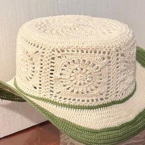 May include: Cream-colored crochet hat with a wide brim and a square pattern design. The brim is trimmed with a green border. This hat is a stylish accessory for sun protection.