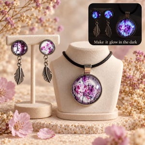 May include: A jewelry set featuring a pendant necklace and two drop earrings. Each piece has a circular glass cabochon with a purple and white floral print and a silver-toned metal feather charm. The necklace has a black cord. Text reads: Make it glow in the dark.